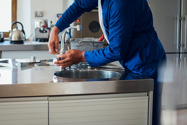 A plumber in a blue uniform performing emergency restoration on the faucet at a kitchen sink, with modern appliances visible in the background.