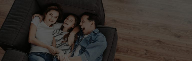 A happy family of three, a mother, father, and young daughter, lying closely together on a dark sofa, smiling and looking at each other with an emergency restoration completed wooden floor background.