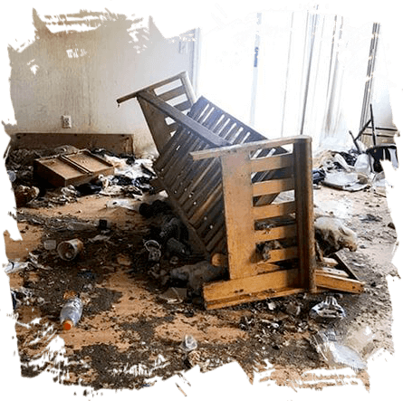 A damaged room with debris scattered all around, awaiting emergency restoration. An overturned wooden bed frame is the focal point, surrounded by broken furniture and litter on a dusty floor.