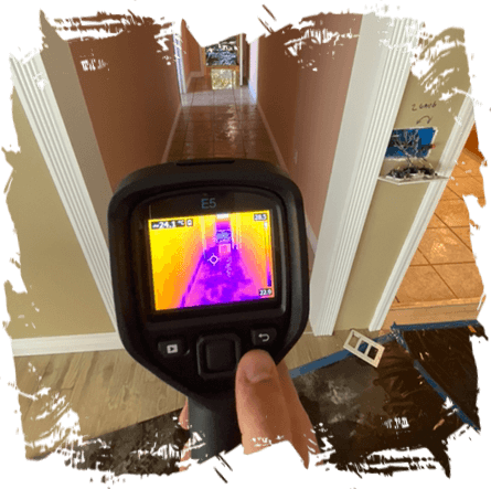 A hand holding a thermal imaging camera during an emergency restoration, with its display screen showing a colorful heat map of a hallway inside a house.