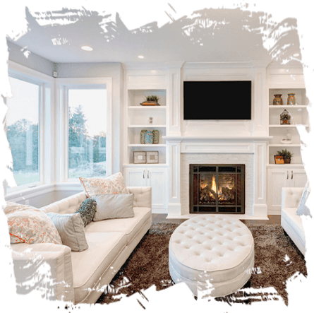 A cozy living room featuring an elegant white fireplace with a mounted TV above, surrounded by built-in shelves, two plush sofas, and a large tufted ottoman, all bathed in natural light