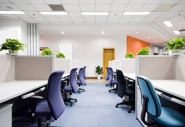 An empty modern office space featuring rows of desks with blue and purple chairs, partitioned by gray dividers, decorated with potted plants, under bright ceiling lights, ready for emergency restoration.