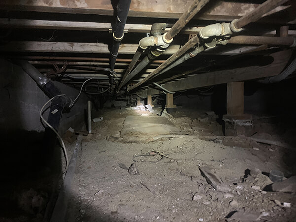 A dimly lit crawlspace under a house with exposed pipes and wooden beams, featuring a beam of light shining towards the center over uneven dirt ground, awaits emergency restoration.