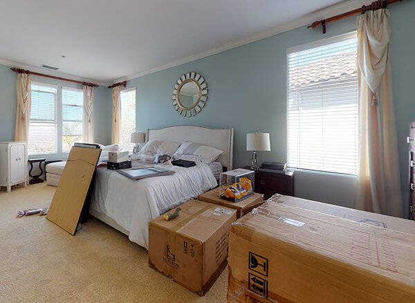 A spacious bedroom with light blue walls and a large bed serves as the site for emergency restoration, cluttered with unpacked cardboard moving boxes and various items spread around, suggesting a remediation in process.