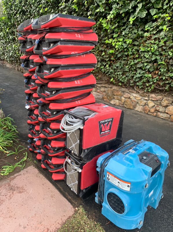 A tall stack of red pizza delivery bags next to large blue and black emergency restoration cooling units placed on a pavement by a green hedge.