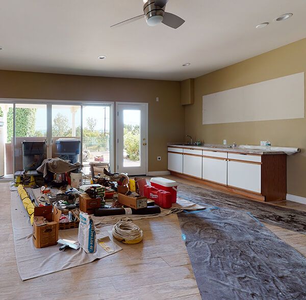 A cluttered living room during emergency restoration with tools, boxes, and belongings scattered on the floor. A modern kitchen is partially visible in the background.