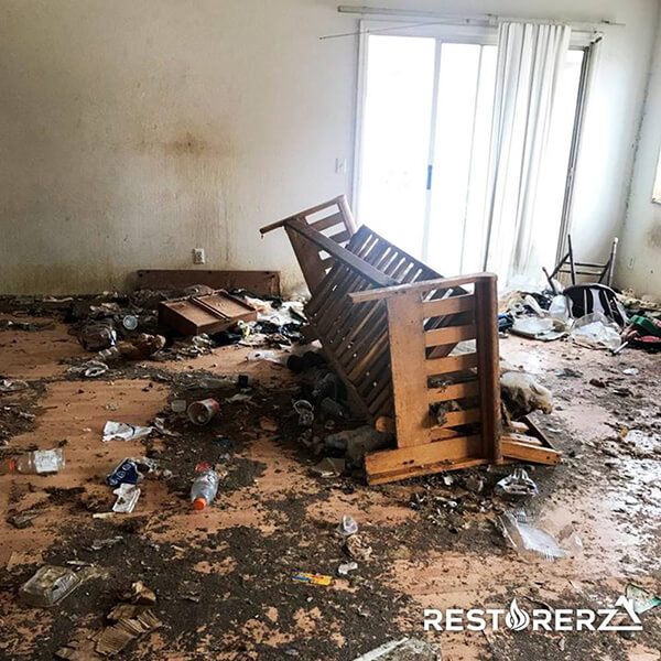 A cluttered, abandoned room with trash scattered all over the floor and a flipped wooden chair in the center. The walls are damp and the paint is peeling, indicating a need for emergency restoration.