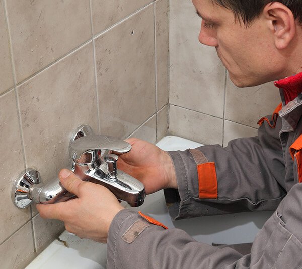 A plumber in a grey and orange uniform performs an emergency restoration, installing a shiny new silver faucet on a tiled bathroom wall, focusing intently on his work.